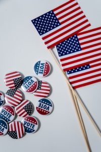 Photo of American flag and vote sticker. Photo by cottonbro: https://www.pexels.com/photo/american-flags-and-pins-on-white-background-4669109/